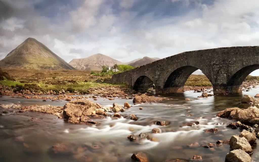 The stone arches of Sligachan Bridge over a rocky river, with hills and mountains in the background on the Isle of Skye.