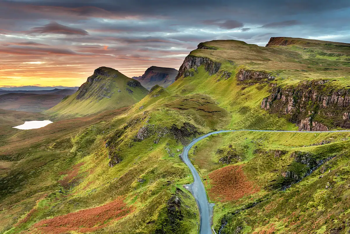 Winding road through dramatic green hills and cliffs under a colorful sky on the Isle of Skye, Scotland.