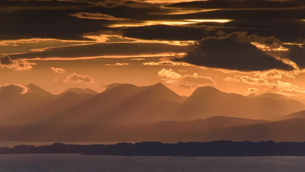 Golden sunset light streaming over dark mountain silhouettes and water on the Isle of Skye, Scotland.