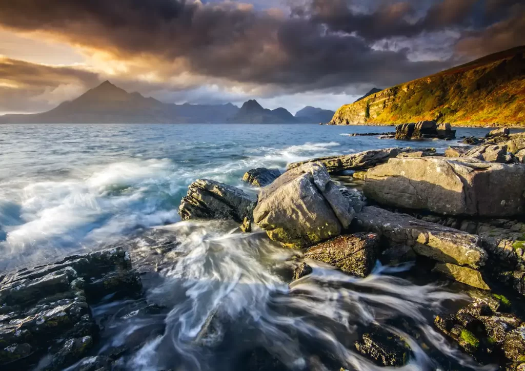 Waves swirling around a rocky shore beneath cliffs and distant mountains at sunset on the Isle of Skye, Scotland.
