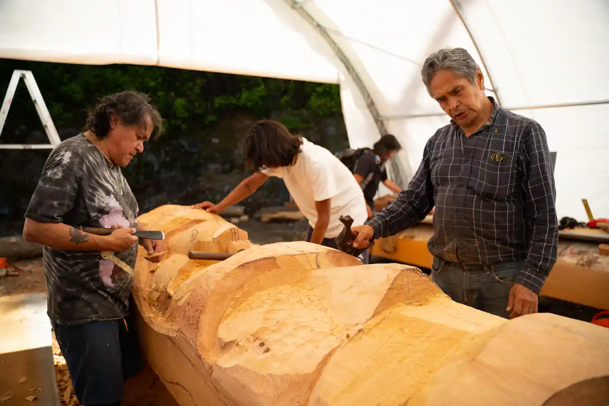 Indigenous woodcarvers shaping a large wooden totem pole beneath a workshop canopy in British Columbia. Photo by Jeremy Williams, courtesy of IISAAK OLAM Foundation.